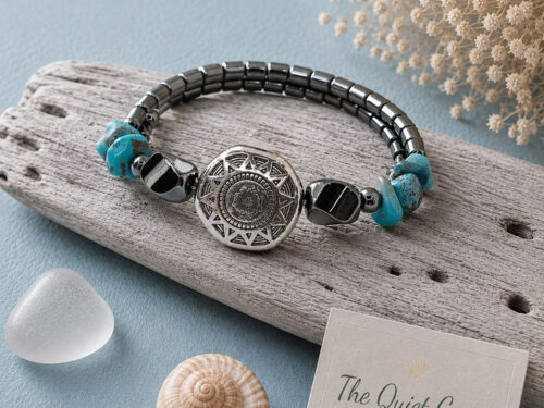 Small Southwestern style memory wire bracelet with polished hematite beads, turquoise chip beads, and a silver tone floral focal bead displayed on a white jewelry stand.