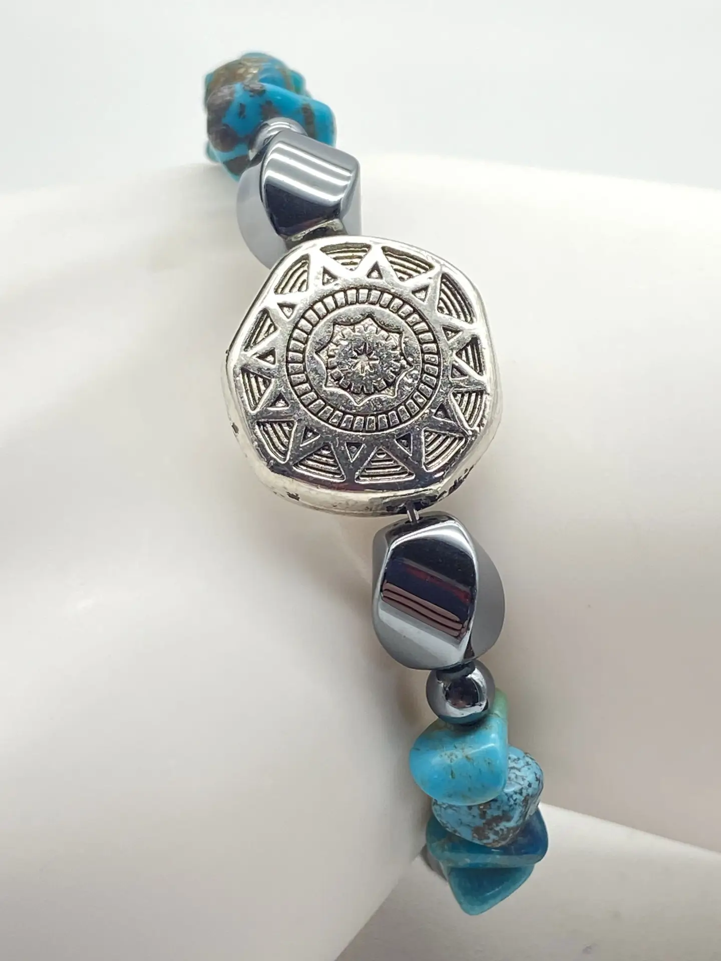 Small Southwestern style memory wire bracelet with polished hematite beads, turquoise chip beads, and a silver tone floral focal bead displayed on a white jewelry stand.