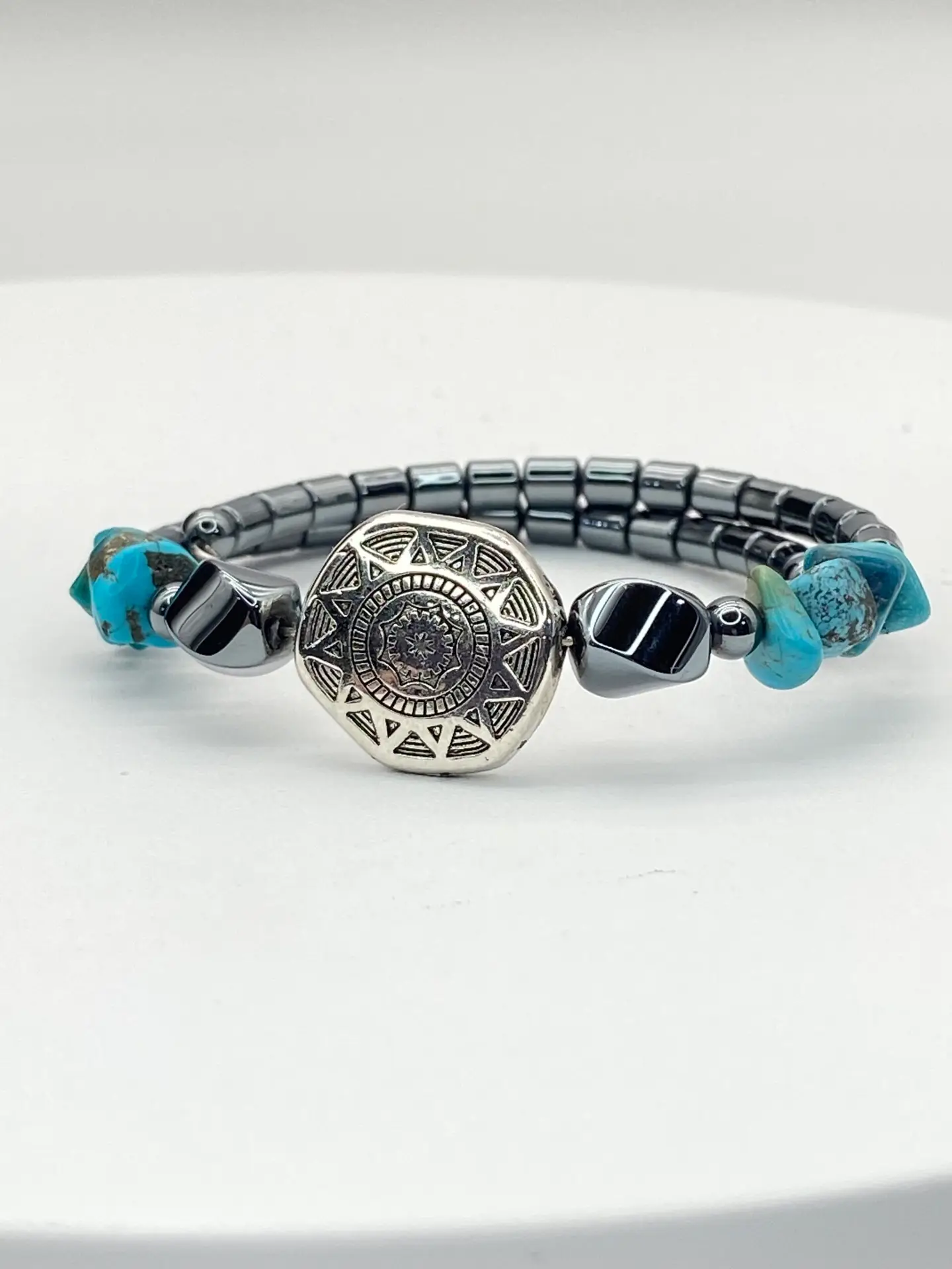 Small Southwestern style memory wire bracelet with polished hematite beads, turquoise chip beads, and a silver tone floral focal bead displayed on a white jewelry stand.