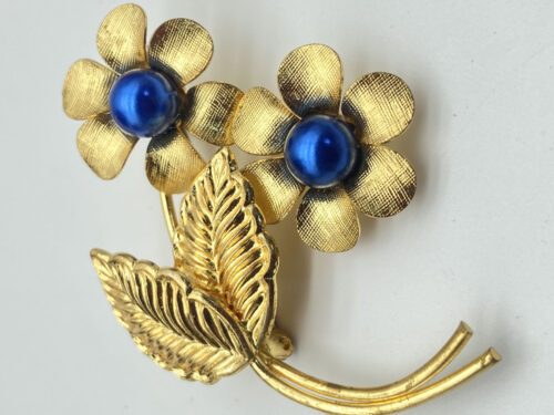Vintage gold-tone floral brooch with two textured flowers, faux blue glass pearl centers, sculpted leaves, and curved stems.